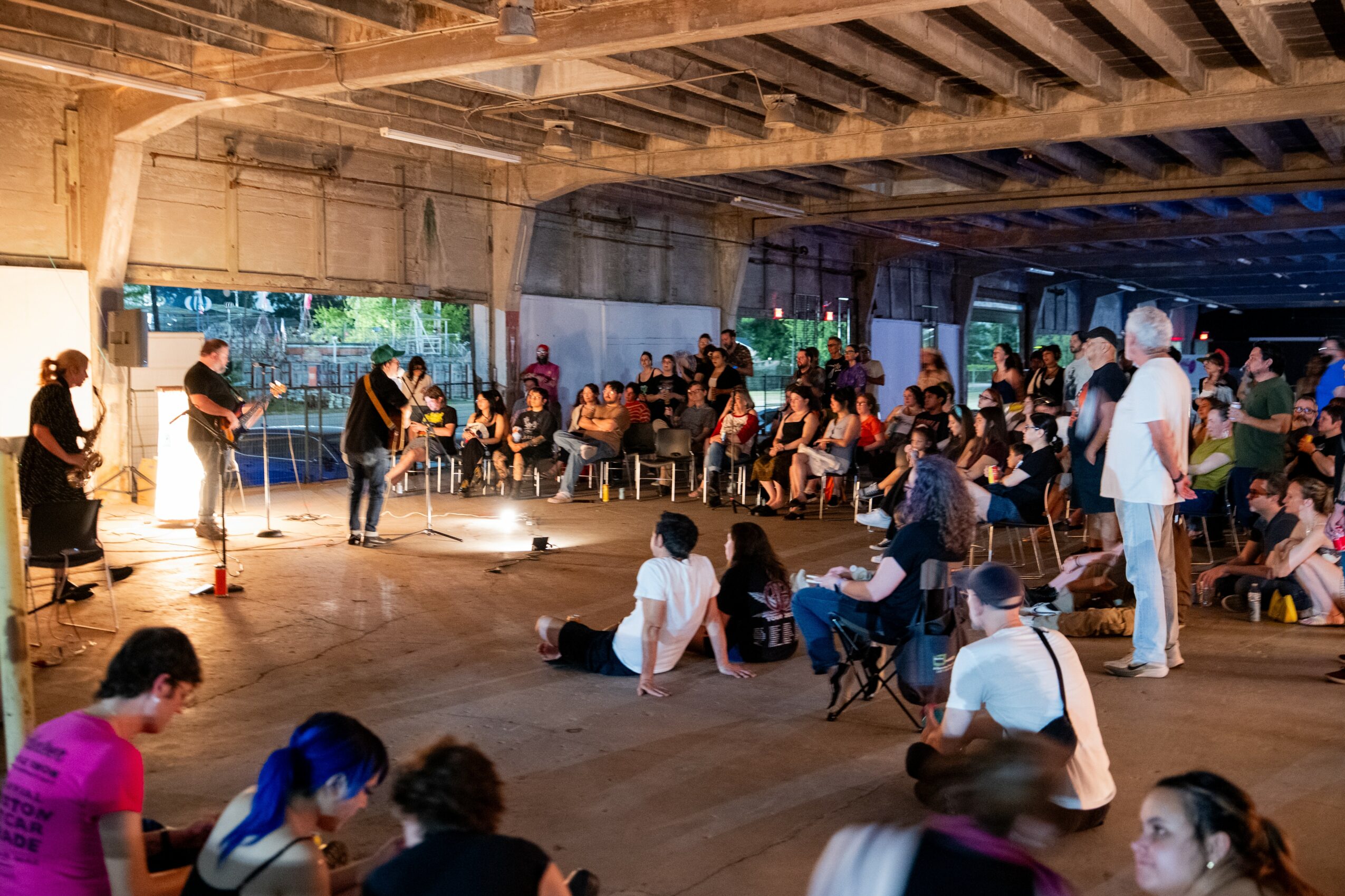 People gather under a bridge, some sitting and some standing, with a performance or event happening in the background.