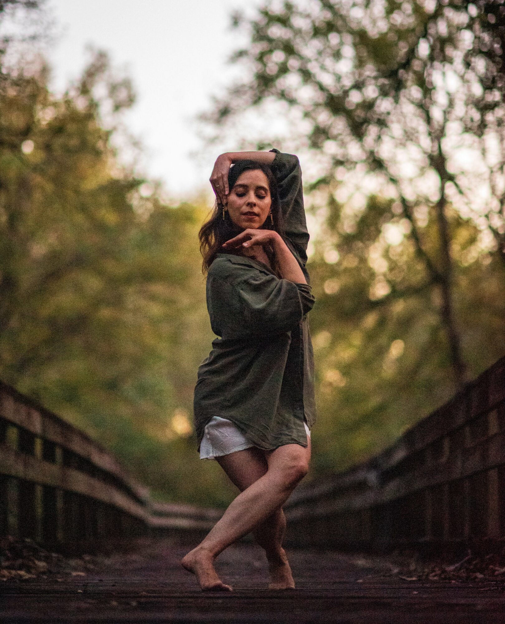 Young woman standing barefoot on a wooden bridge surrounded by trees, with one hand on her head and the other on her shoulder.