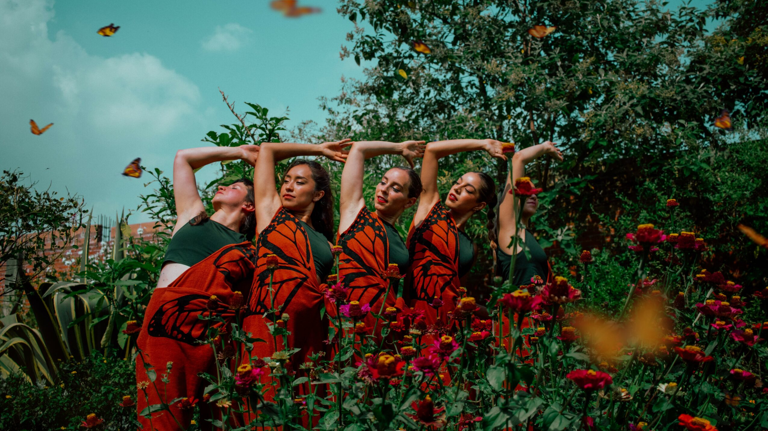 Four women in red and black dresses pose with arms above heads in a garden with flowers and butterflies.
