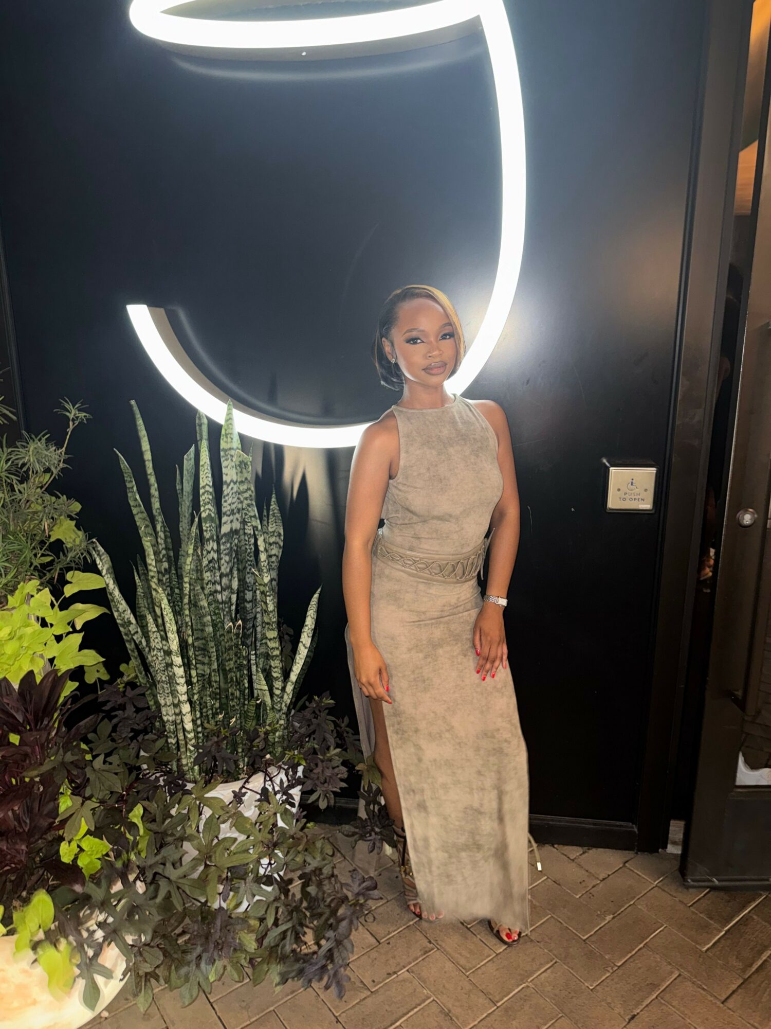 Woman in a long, light-colored dress standing indoors near plants and a black wall with a glowing circular design.