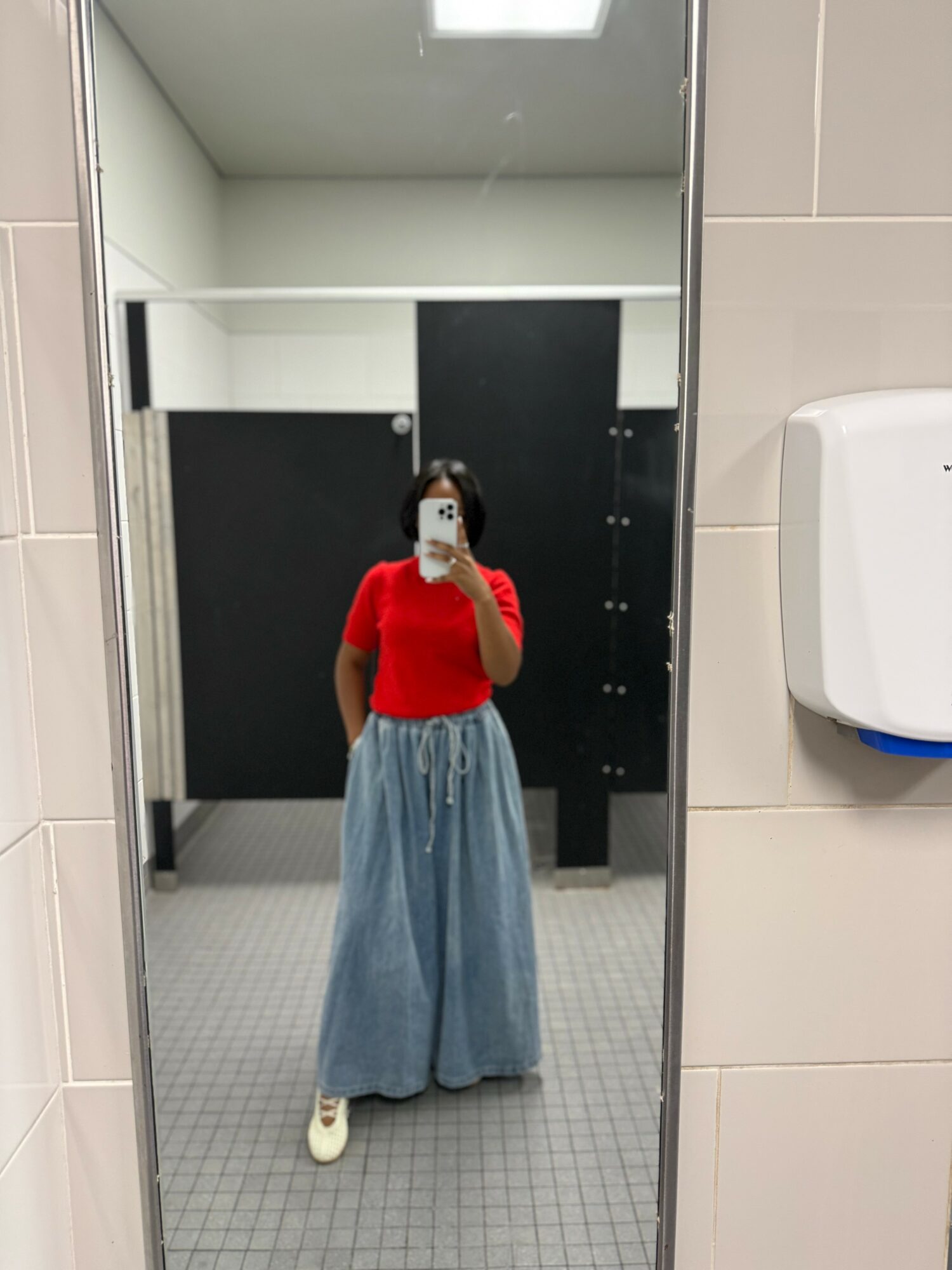 Person taking a mirror selfie in a bathroom, wearing a red top and wide-leg jeans, with a white mask, tiled walls, and a paper towel dispenser.