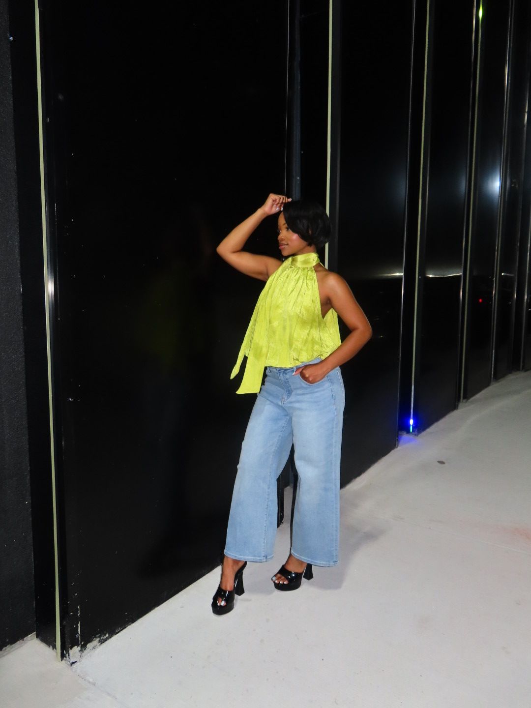 Person standing sideways, wearing a yellow top and light blue jeans, in front of black doors, with a white wall on the right.