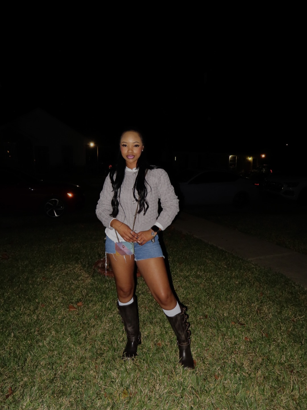 Young woman standing outdoors at night, smiling, wearing a white jacket, denim shorts, and black boots, with a backpack.