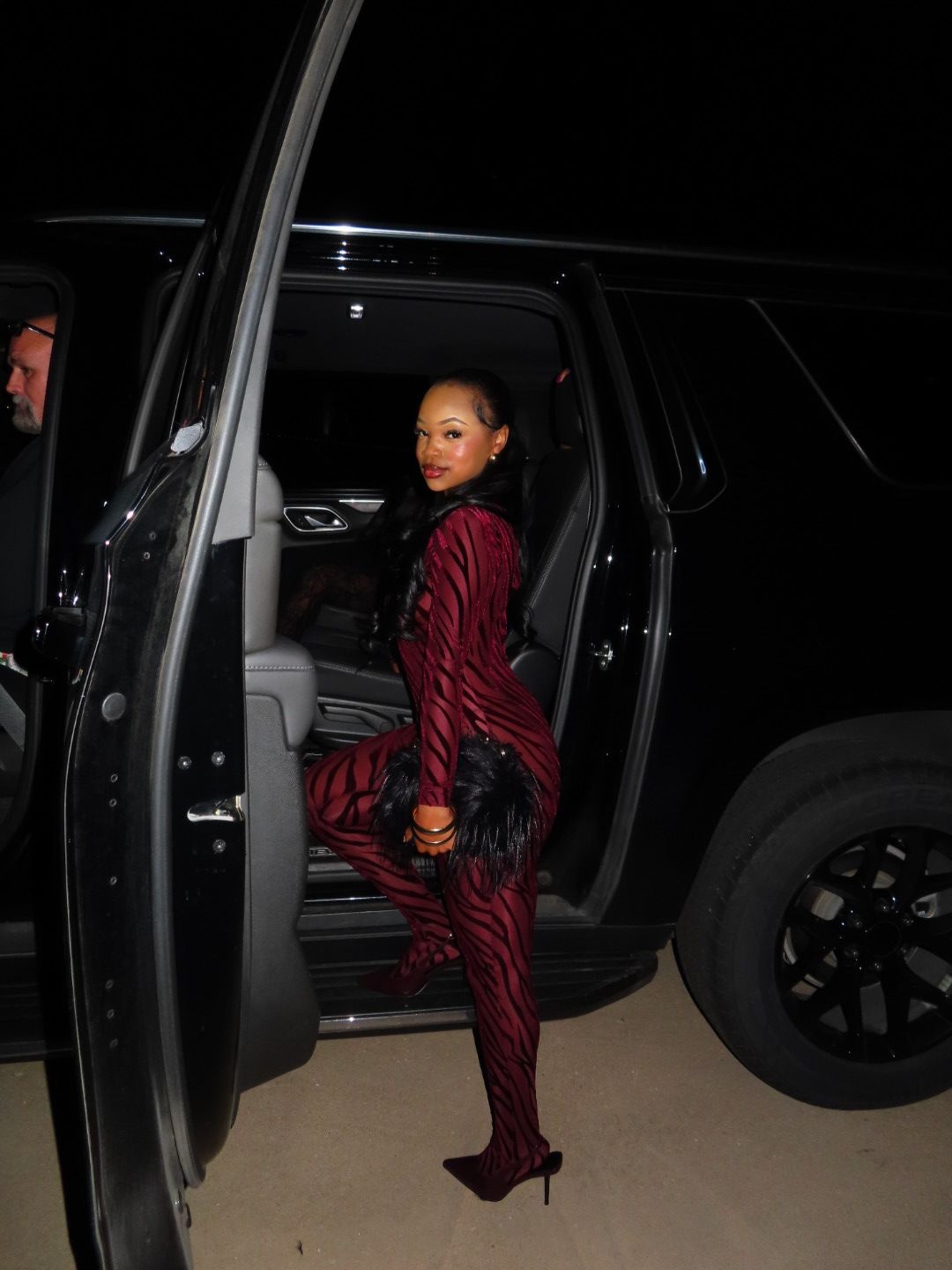 Woman in red striped outfit sitting in a car with open door, holding a clutch, in a garage.