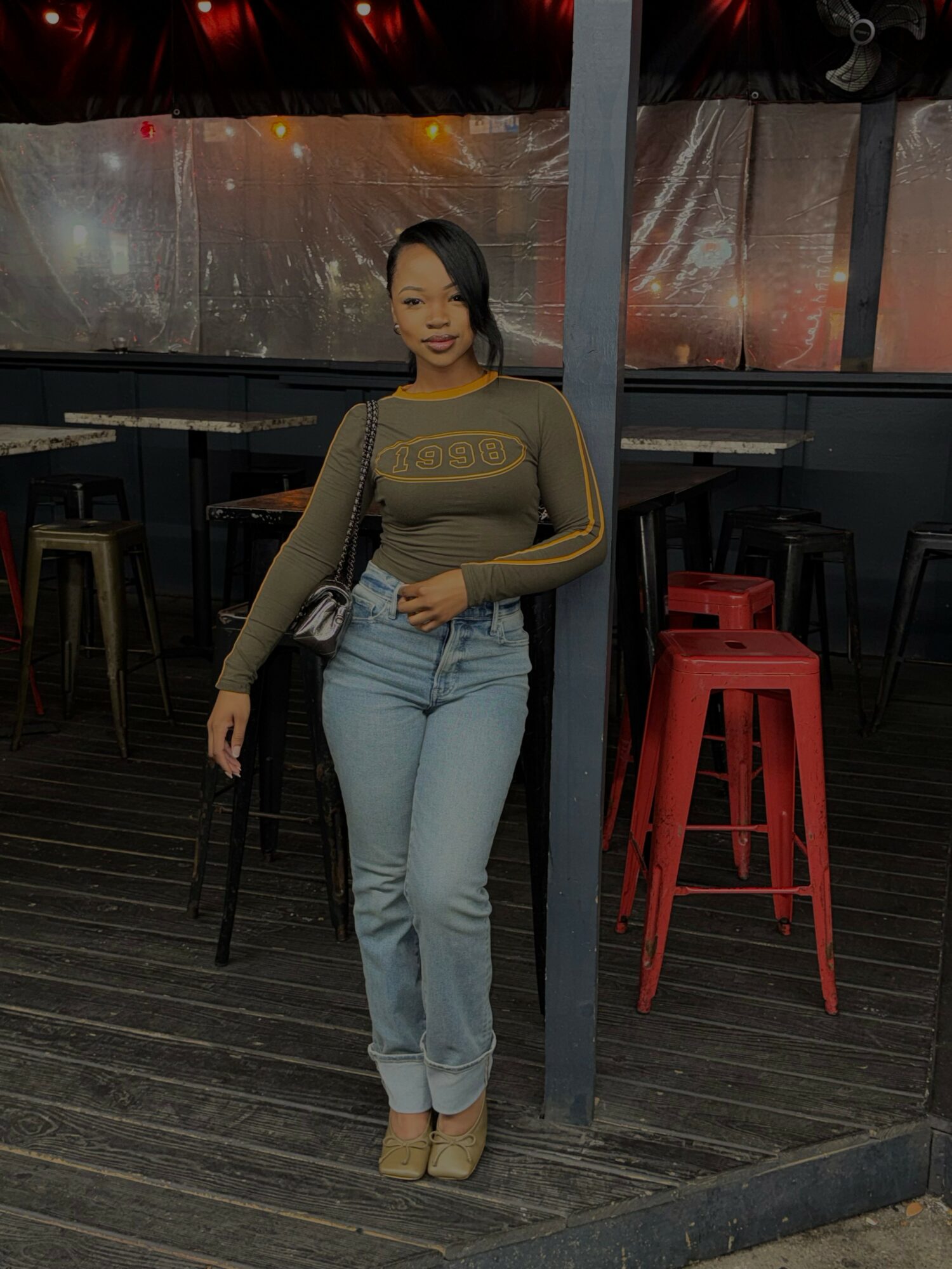 Woman standing indoors with tables and red stools, wearing a long-sleeve top and jeans, smiling.