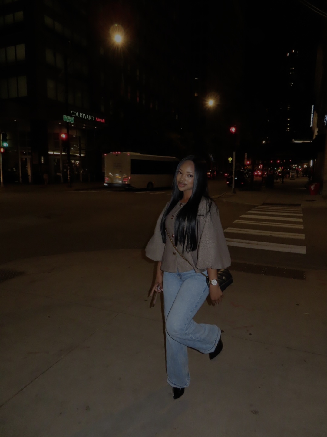 Young woman standing on city street at night, crosswalk visible behind her, illuminated buildings and streetlights in background.