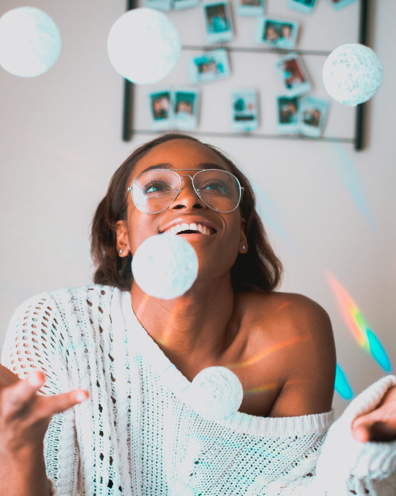 Smiling woman with glasses and a white sweater, surrounded by hanging and falling white spherical decorations.