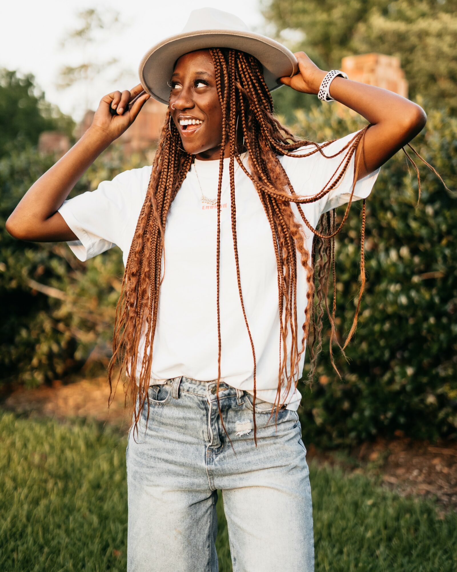 Woman outdoors wearing a hat, smiling, with long braided hair, white t-shirt, and jeans, holding her hat with both hands.