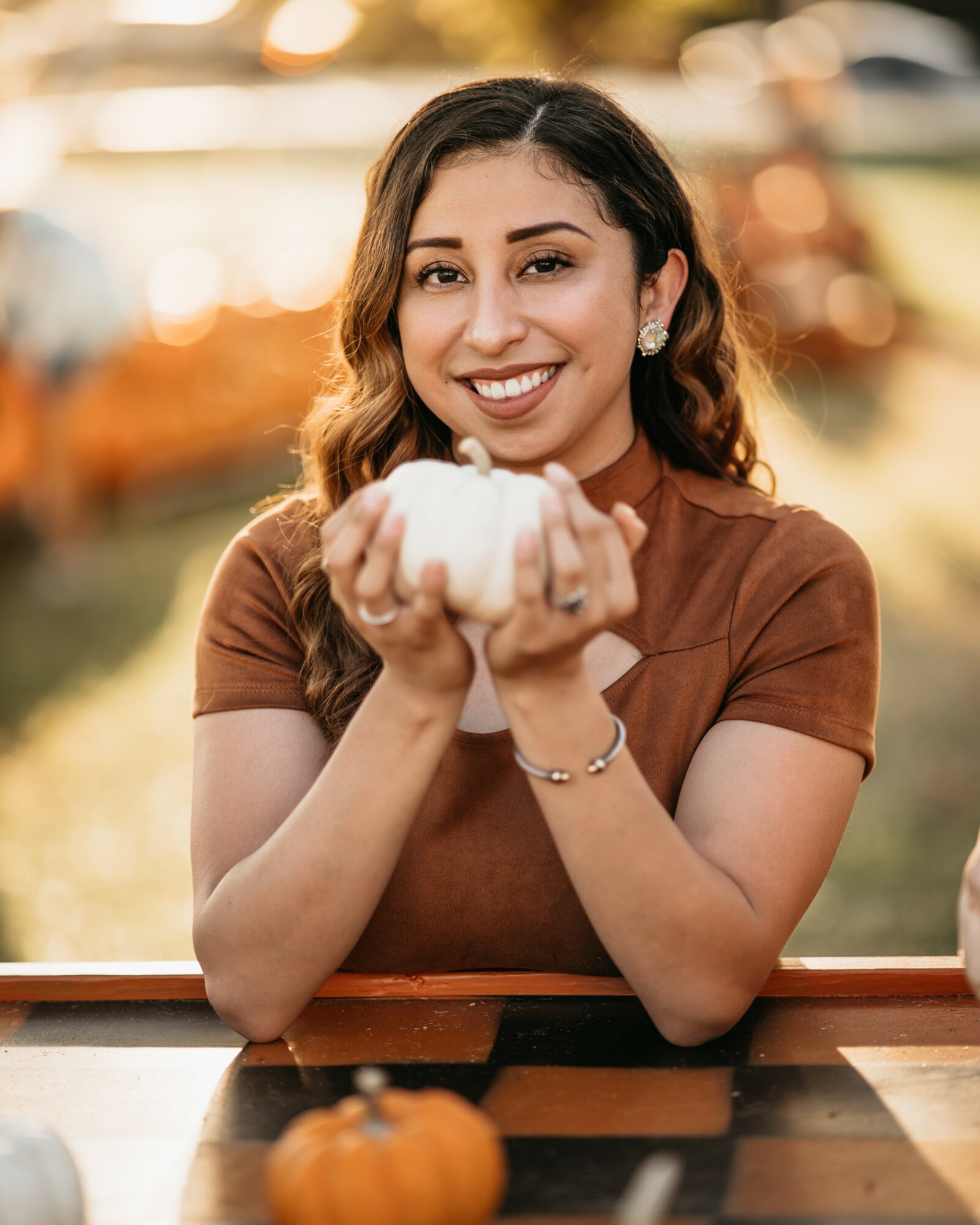 Smiling woman holding a small pumpkin outdoors, with blurred background of trees and cars.