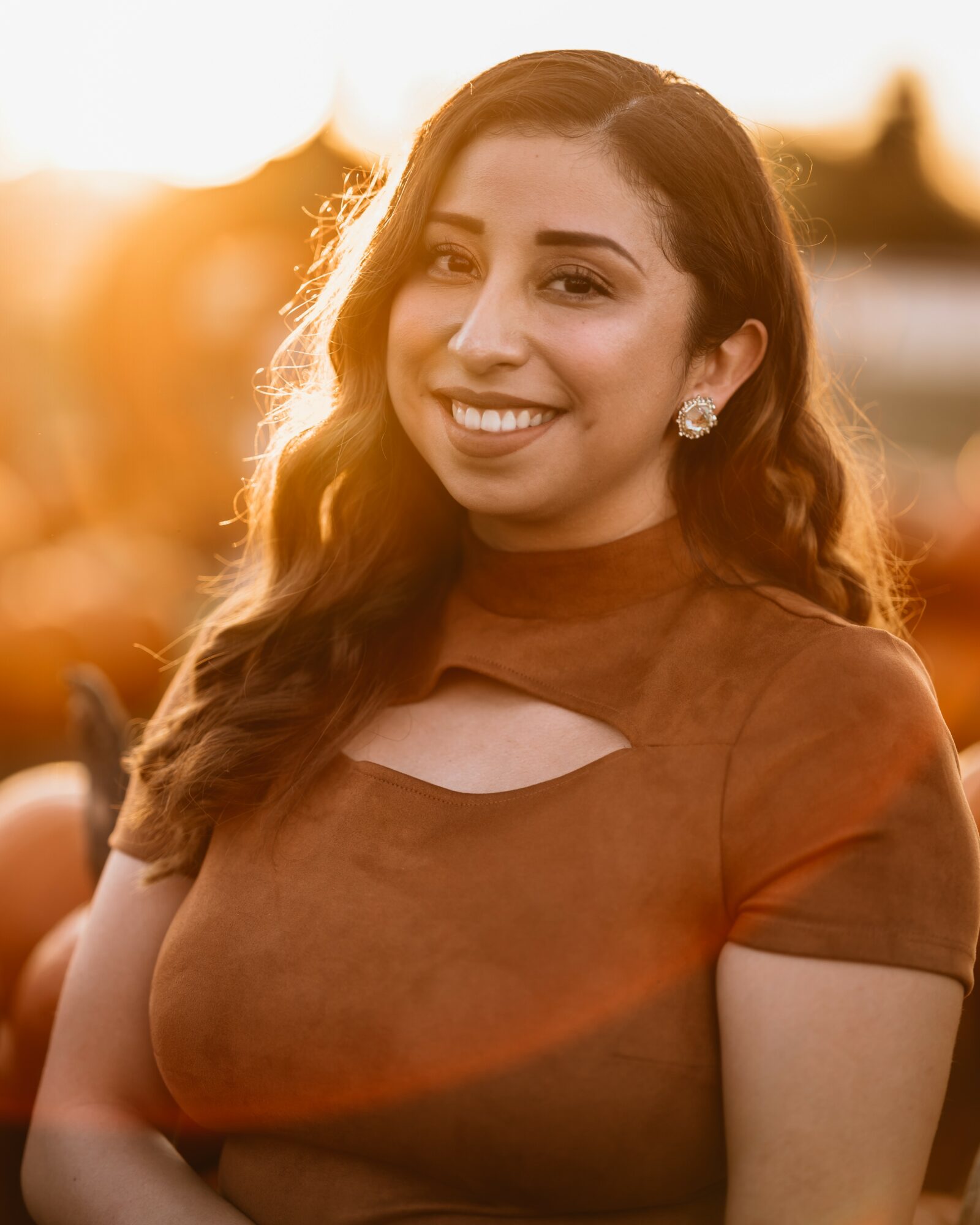 Young woman smiling outdoors during sunset, wearing a brown top with a cutout, with sunlight behind her.