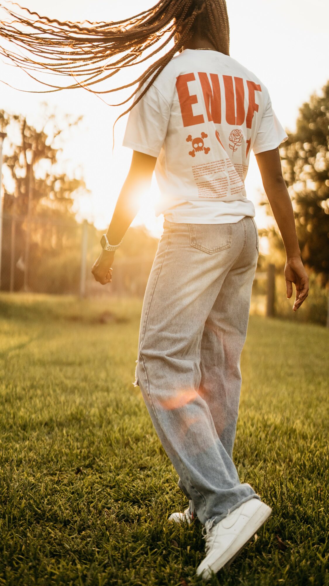Person standing outdoors on grass with sunlight behind, wearing a white t-shirt and light-colored pants, hair flowing.