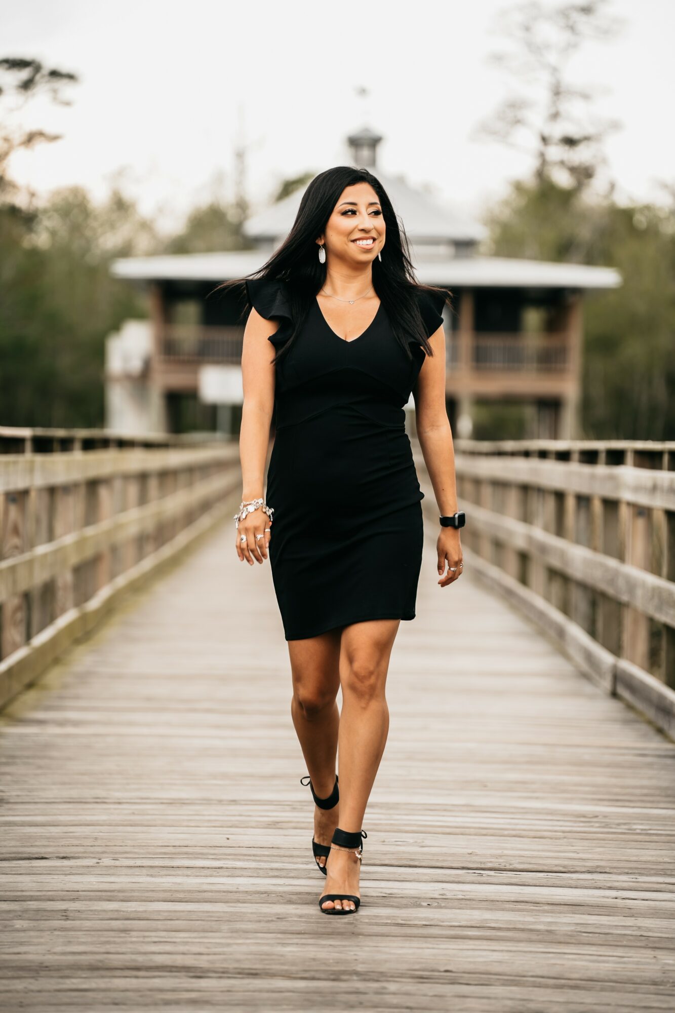 Woman in black dress walking on a wooden bridge outdoors, smiling, with trees and a building in the background.