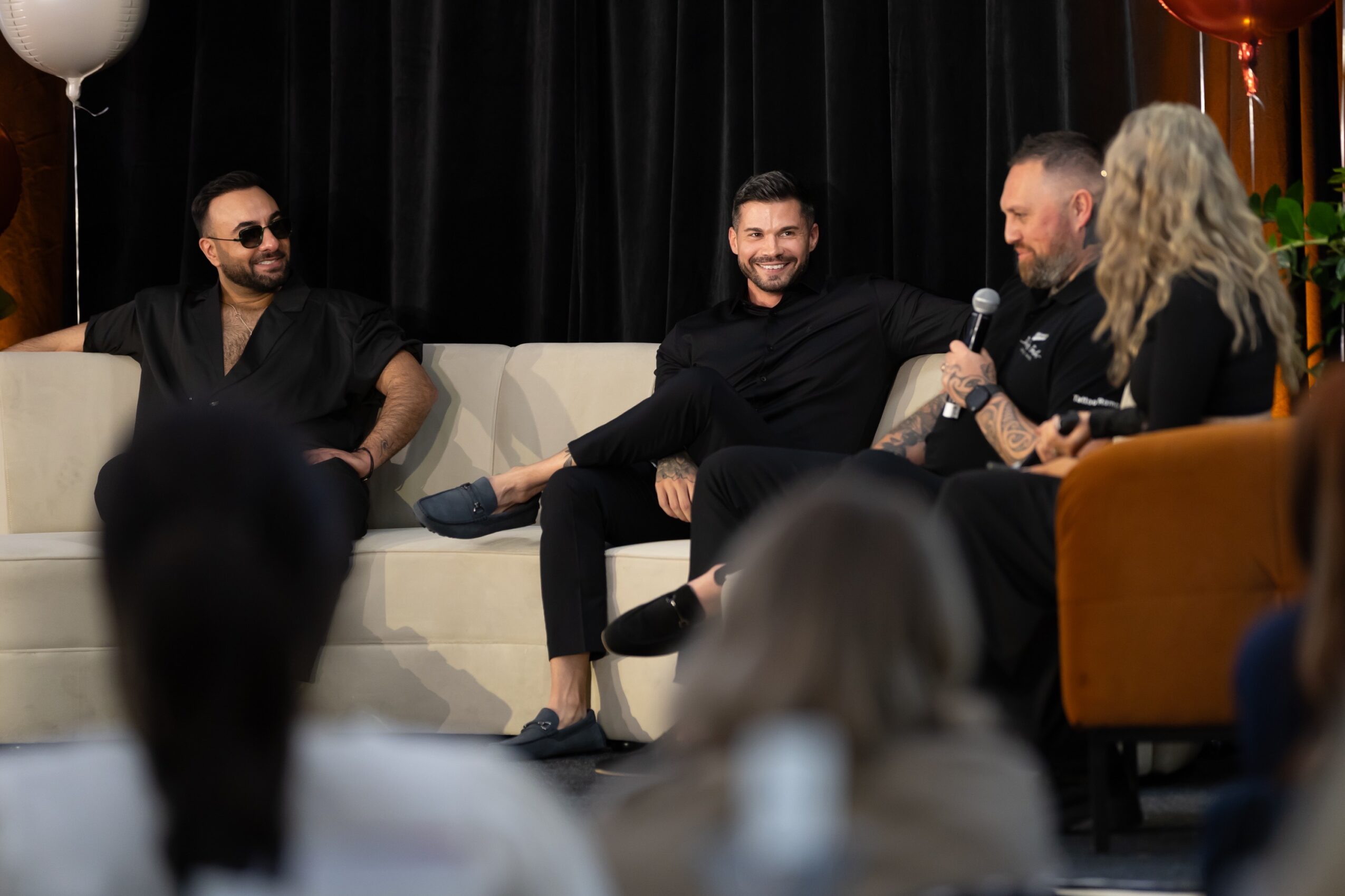 Four people sit on a stage with a black curtain background, engaged in conversation, with an audience in front.