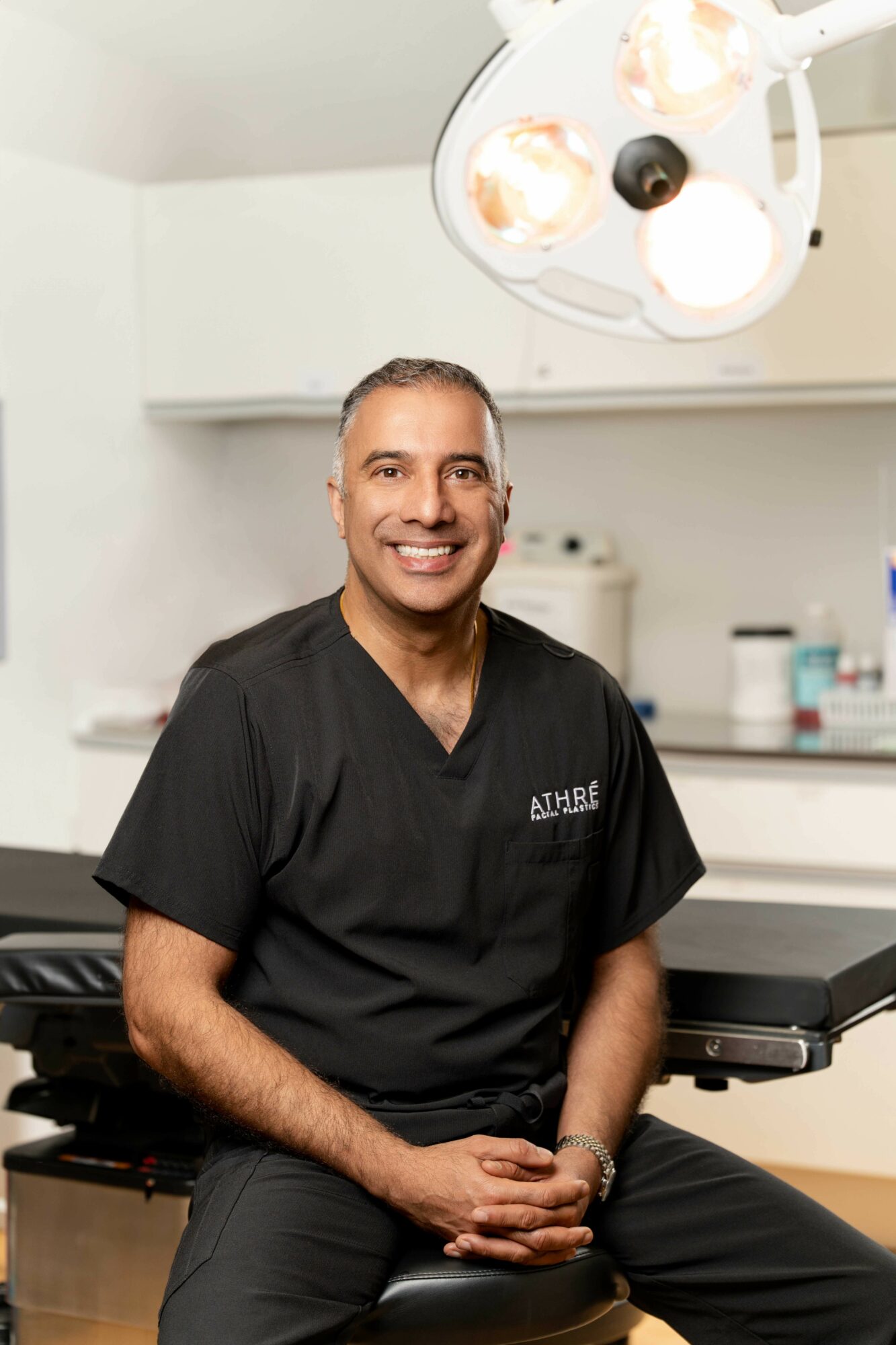 Smiling man in black medical scrubs sitting in a clinical setting with surgical lights overhead.