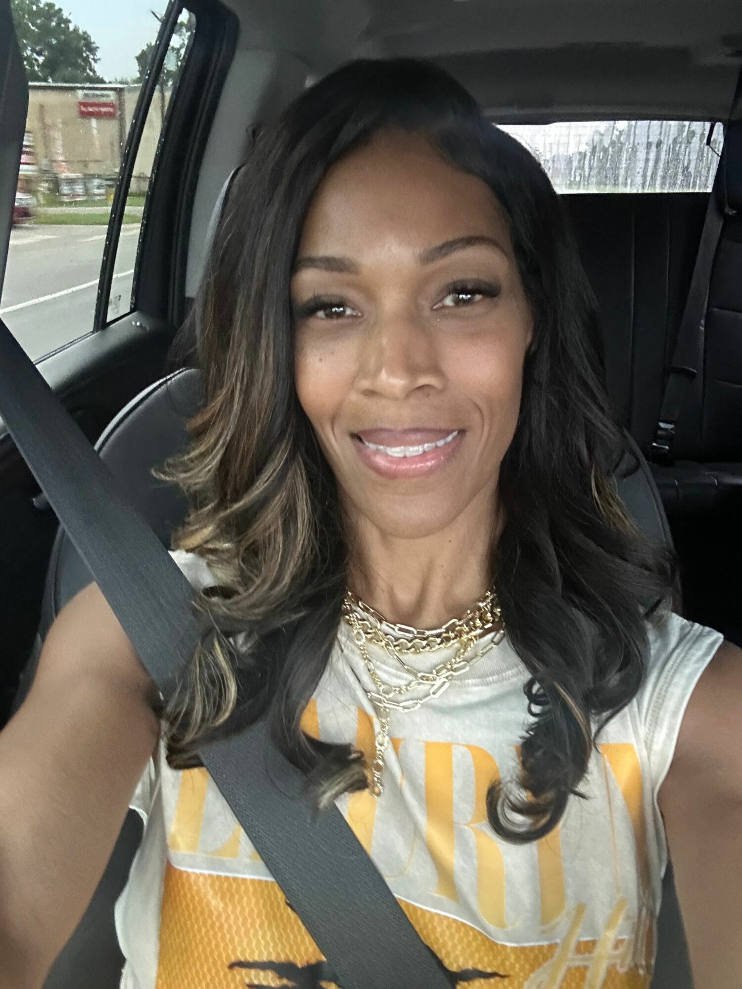 Woman with long dark hair smiling, wearing a gold necklace and a striped shirt, sitting in a car seat.