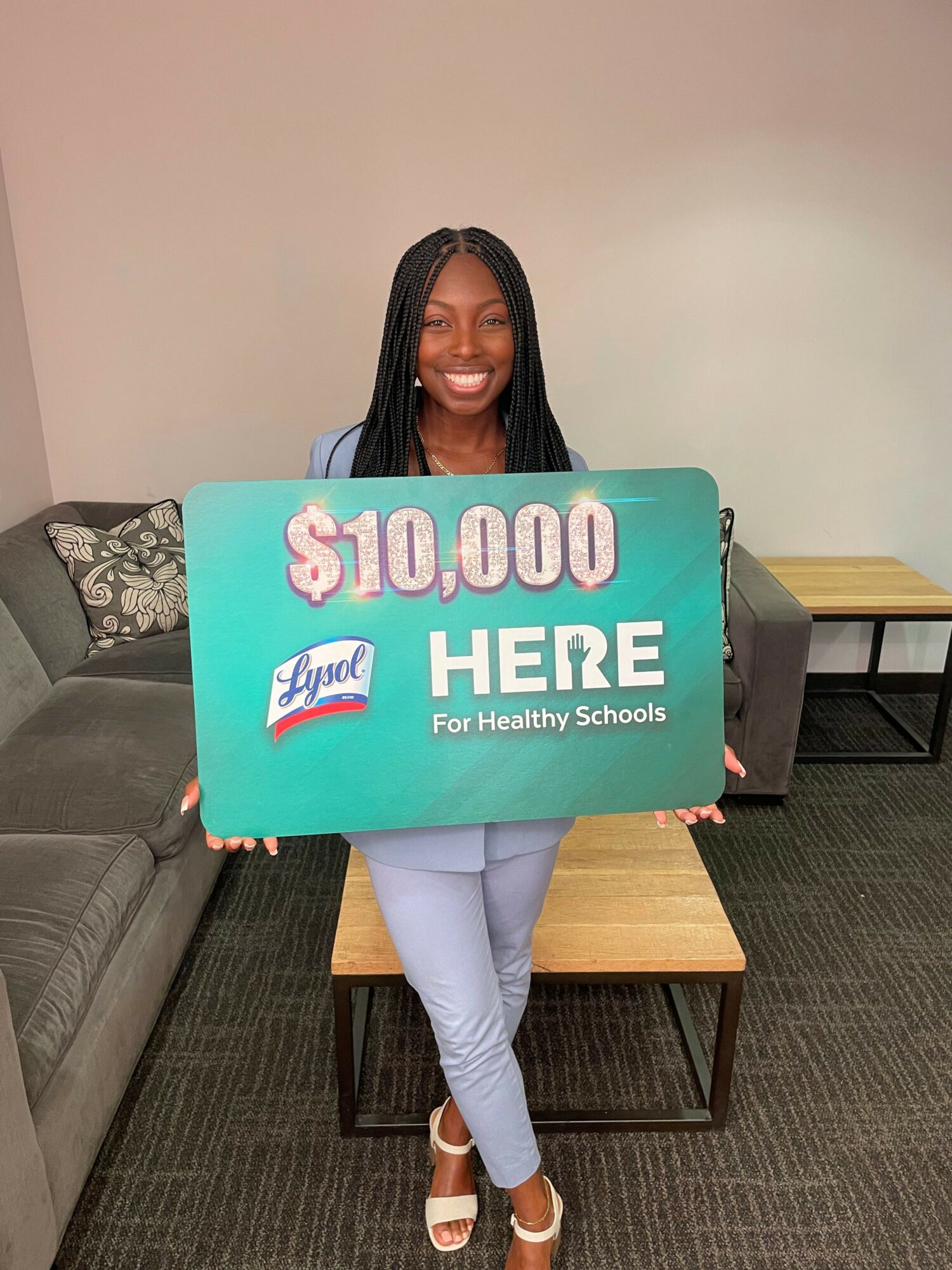 Person holding a large $10,000 check for a scholarship, standing in a room with furniture.