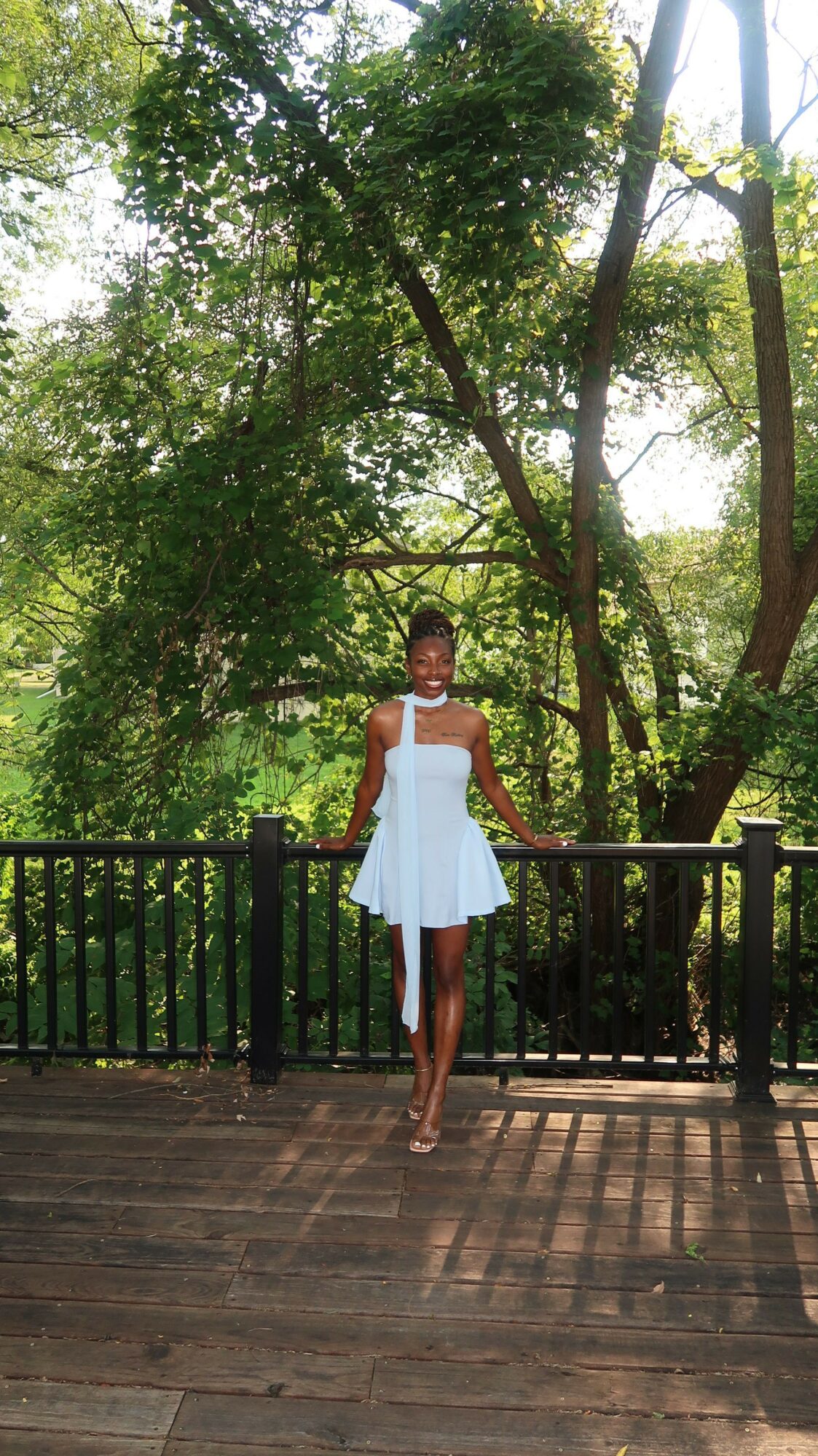 Woman in white dress standing on wooden deck with trees and sunlight in background.