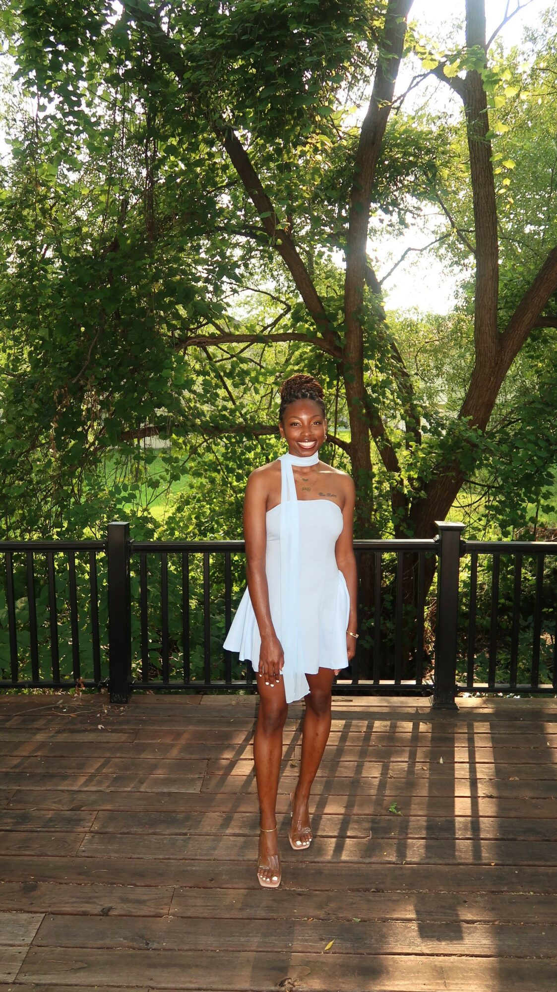 Woman in white dress standing on wooden deck with trees and sunlight in background.