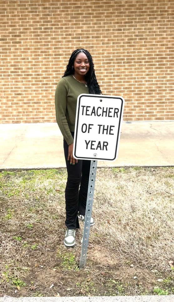 Young woman standing outdoors next to a sign that reads 'Teacher of the Year' in front of a brick wall.