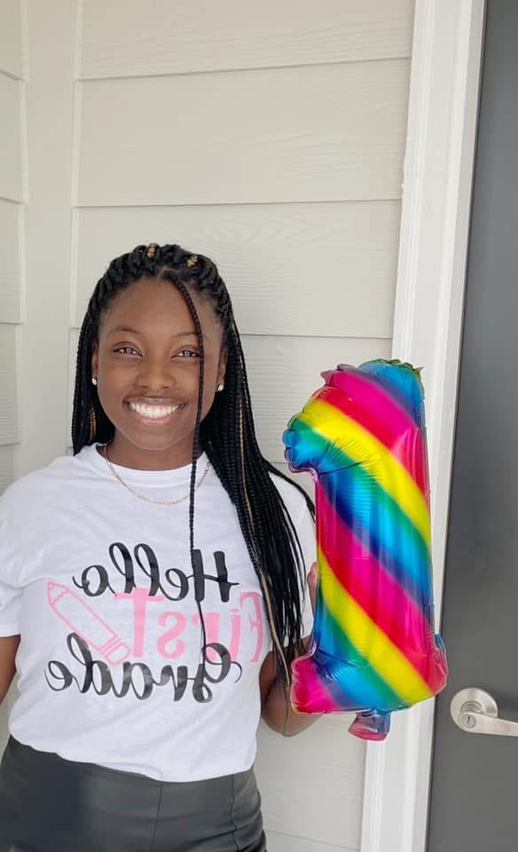 Smiling person with braided hair holding a rainbow-colored balloon, standing outside near a door and wall.