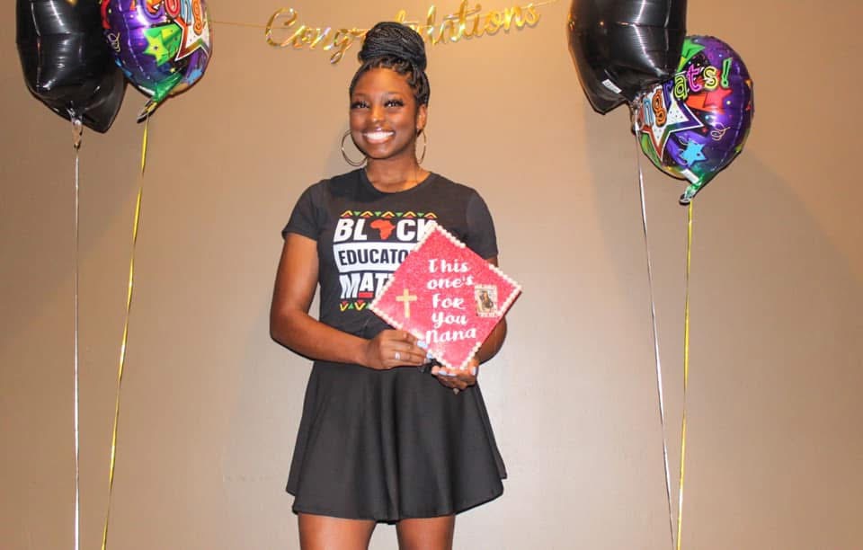 Young woman smiling, holding a red and white sign, standing between black balloons with colorful designs, celebrating graduation.