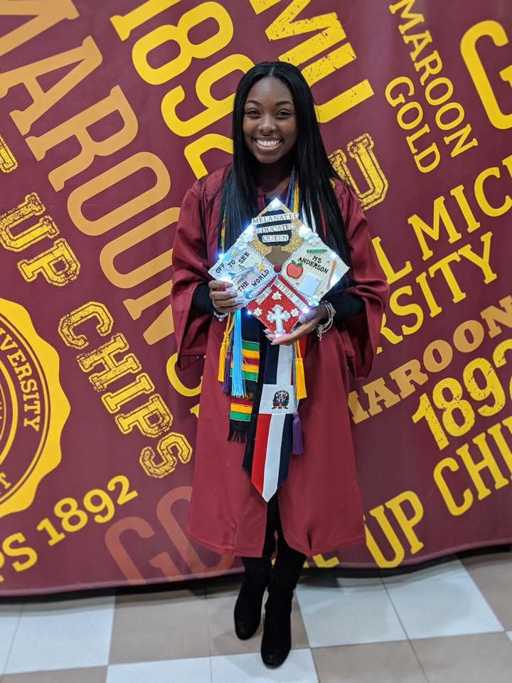 Young woman in graduation gown holding diploma and gift bag, standing in front of university banner, smiling.