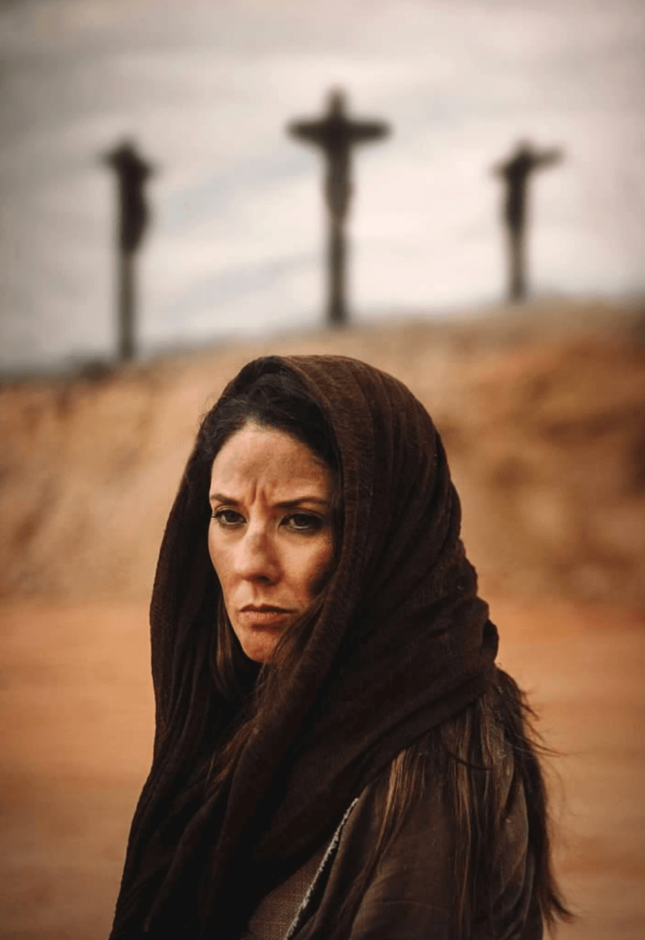 Woman with dark hair and headscarf looking serious, crosses in the background, desert landscape.