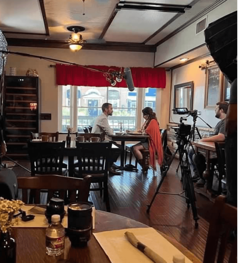 Two people sit at a table in a restaurant, with a camera on a tripod nearby, and a window with red curtains behind them.