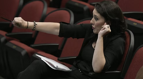 Woman with dark hair sitting in a theater or auditorium, holding a notebook and pointing with her right arm.