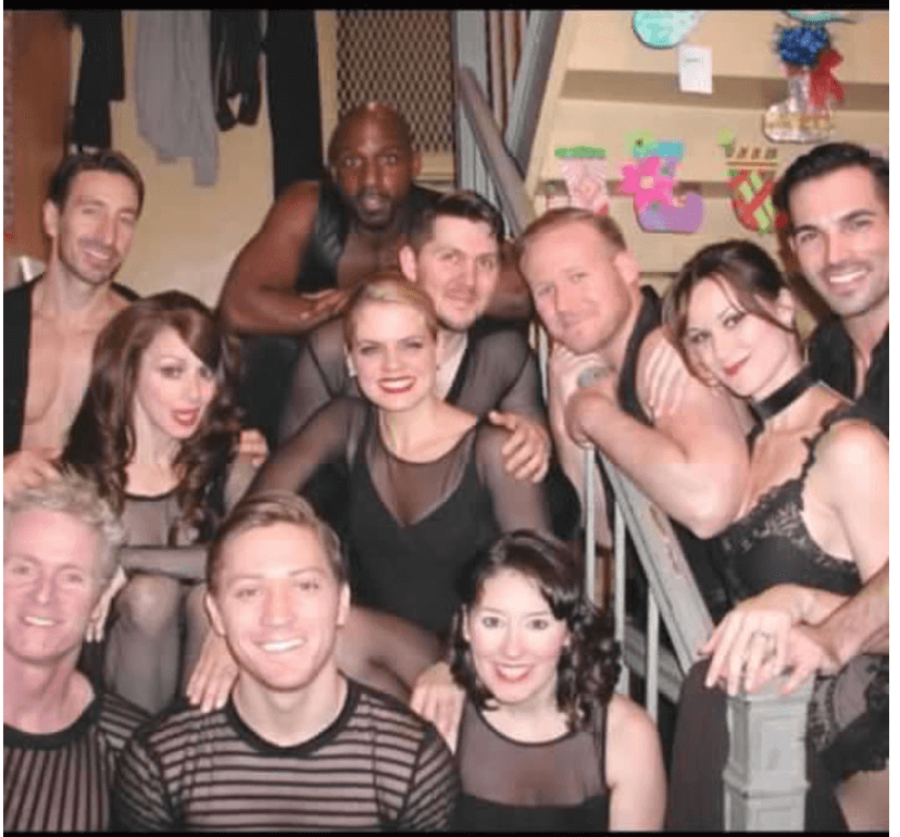 Group of people smiling and posing together indoors, some wearing black clothing, with colorful decorations in the background.