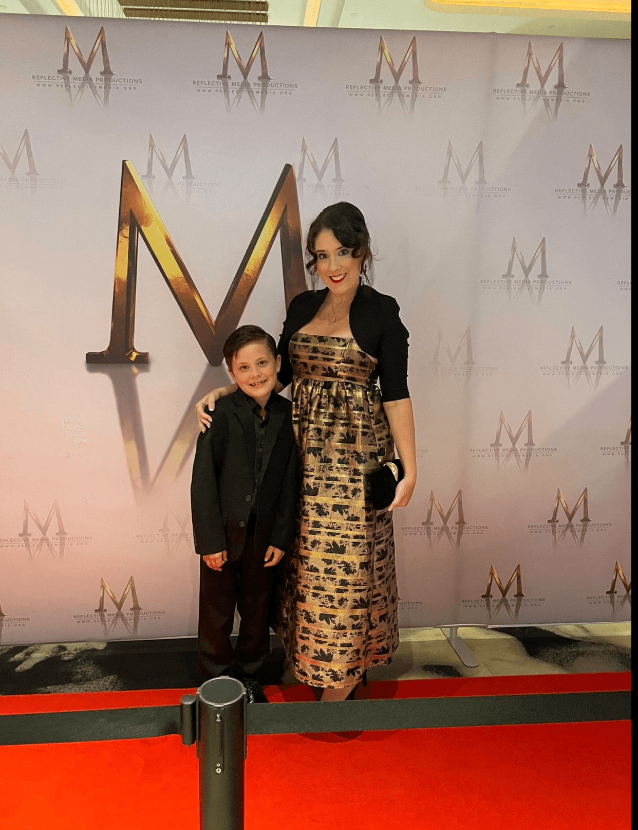 A woman and a young boy stand together on a red carpet in front of a backdrop with a large gold letter M and smaller M logos. The woman wears a long patterned dress and black jacket, and the boy wears a black suit. Both are smiling.