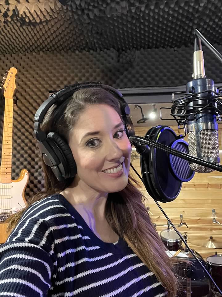 Woman in a recording studio wearing headphones, smiling, with a microphone in front of her, and a guitar in the background.