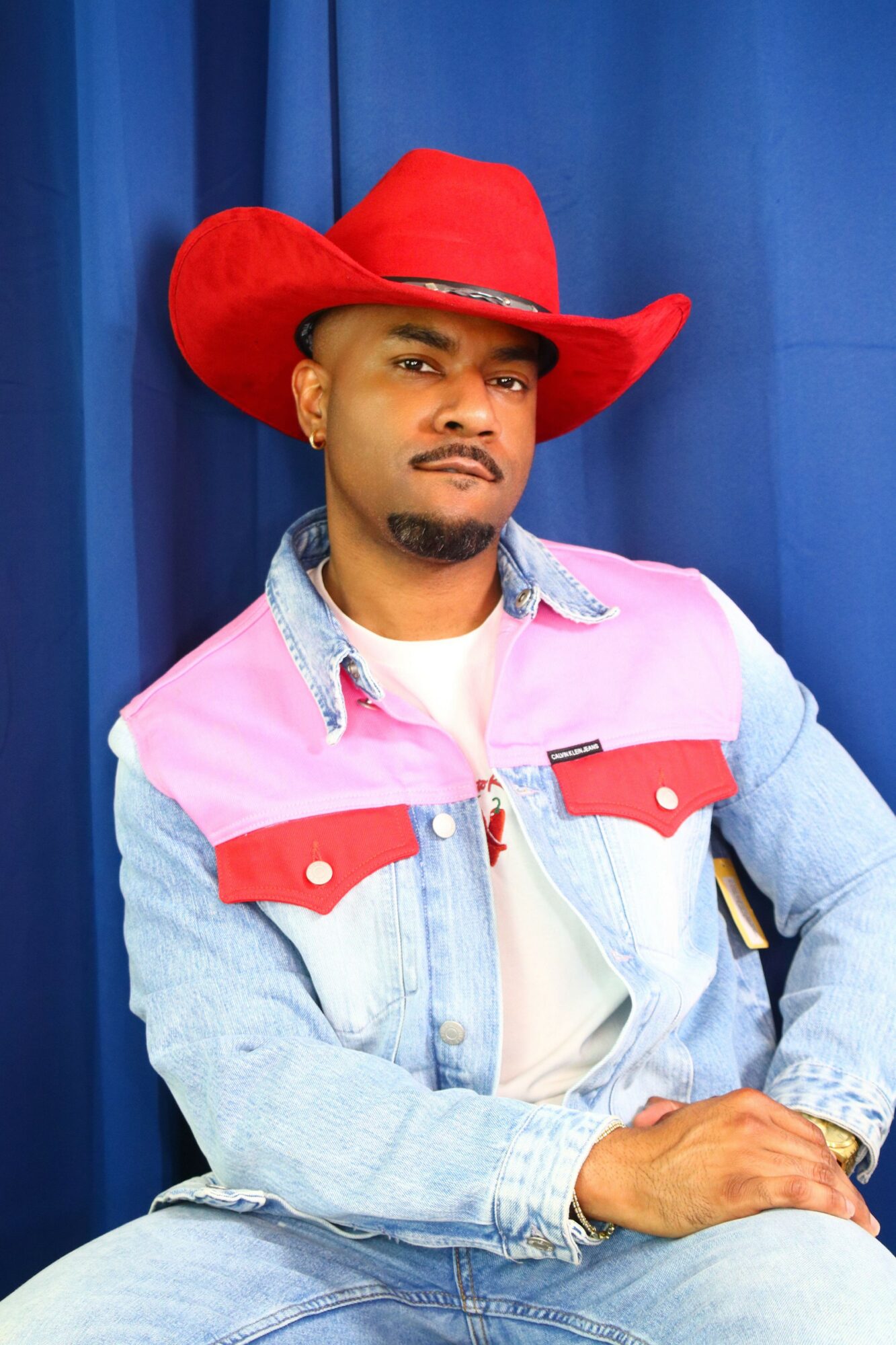 Man wearing a red cowboy hat, denim jacket with pink and red accents, sitting against a blue curtain background.