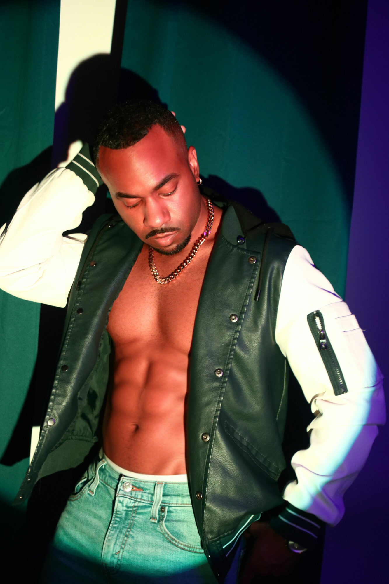 Young man with a muscular torso, wearing a black and white jacket, standing against a colorful background.