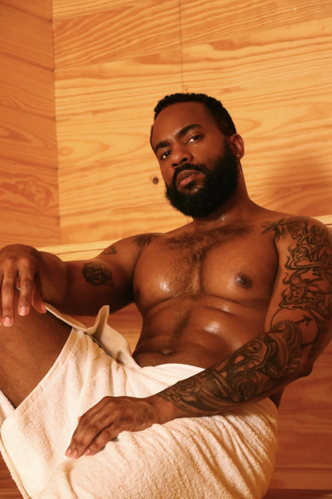 A shirtless man with a beard and tattoos on his arms and chest, sitting against a wooden wall, wearing light-colored shorts.