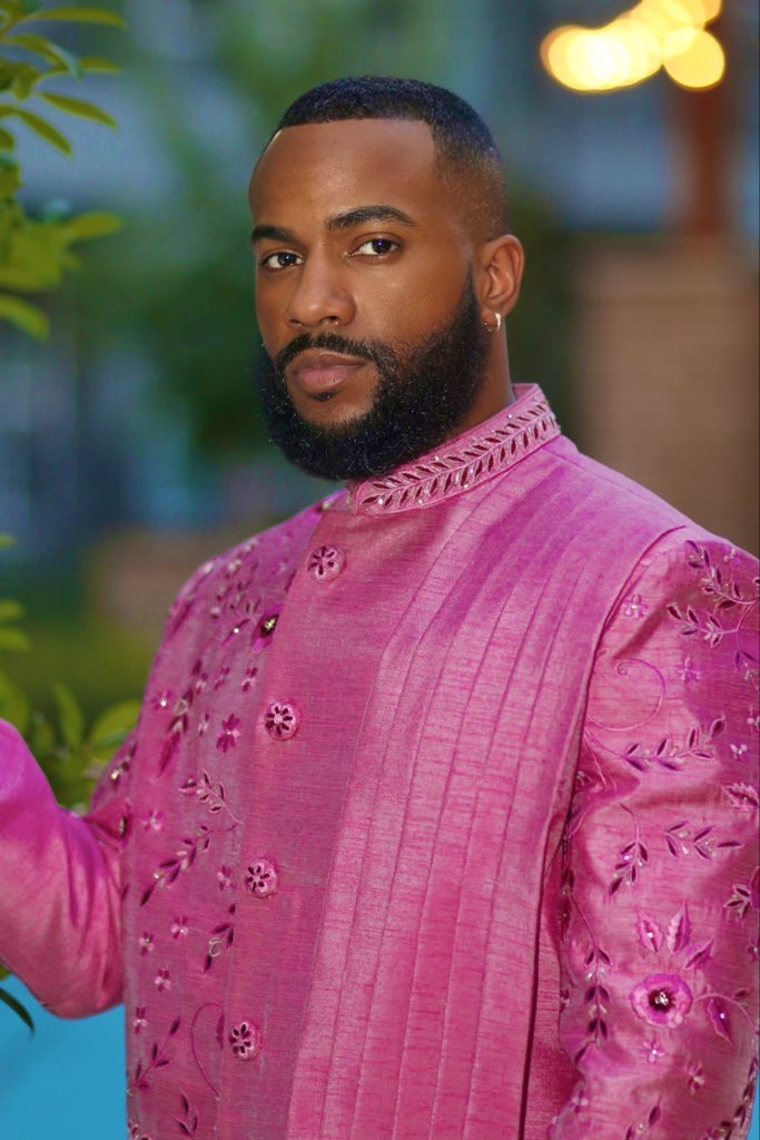 Man with a beard and short hair wearing a pink traditional outfit outdoors, blurred background with greenery and lights.
