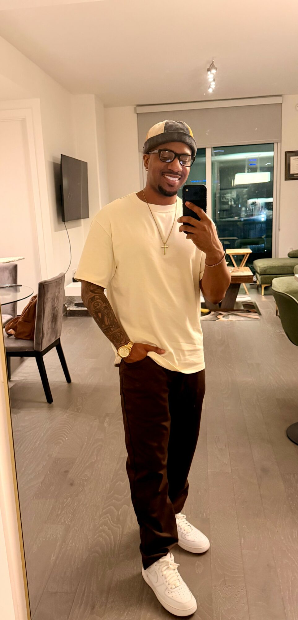 Man taking a mirror selfie in a living room, wearing glasses, a beige t-shirt, dark pants, and white sneakers.