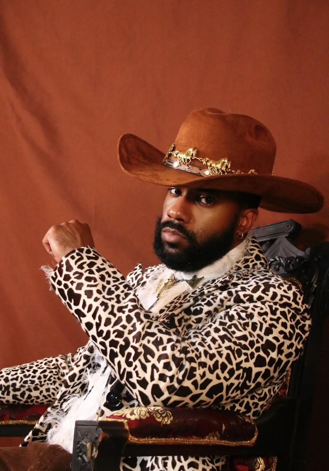 Man wearing a large cowboy hat and leopard print jacket, sitting against a reddish background.