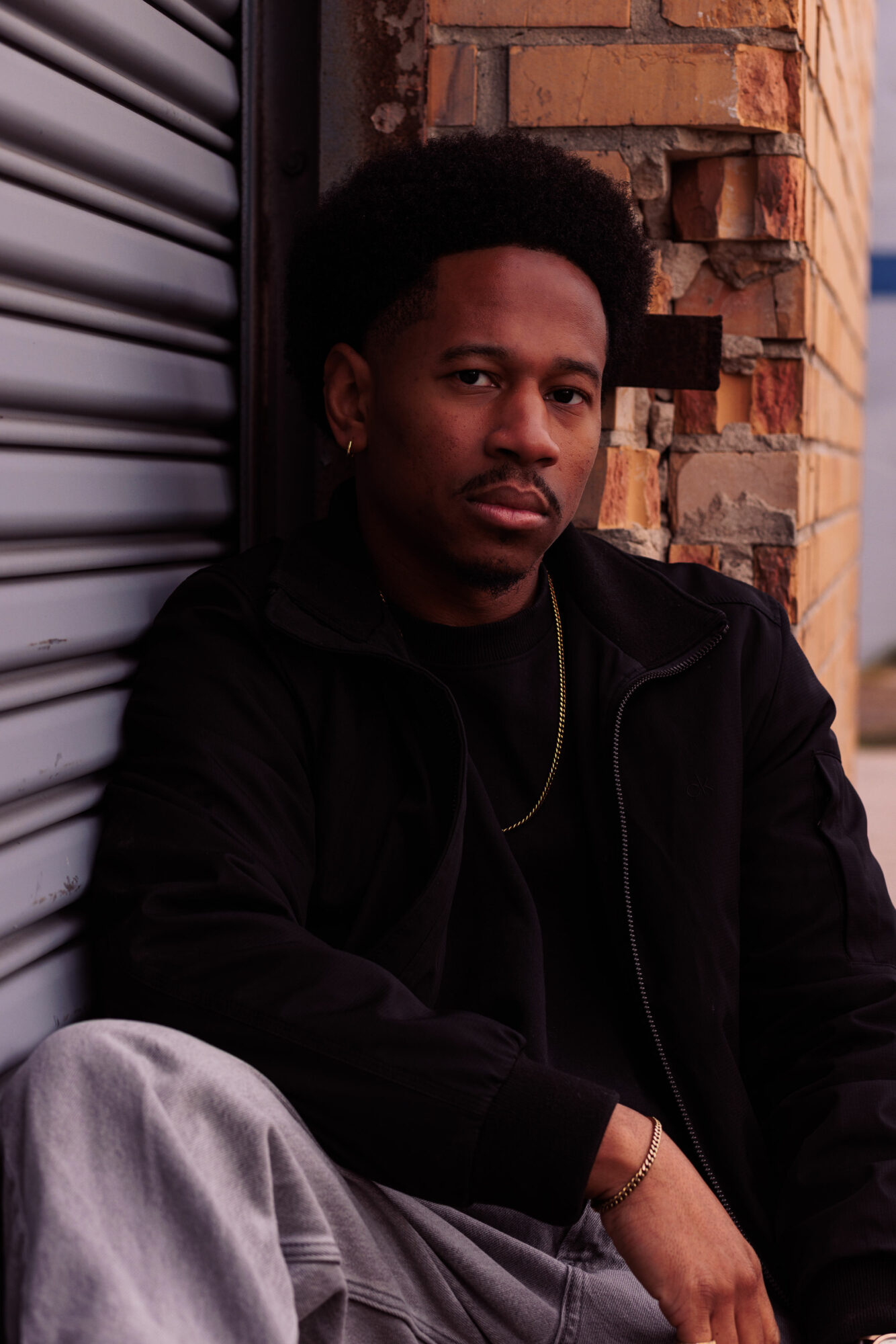 Young man with curly hair and a mustache sitting against a brick wall and metal shutter, wearing dark clothing.