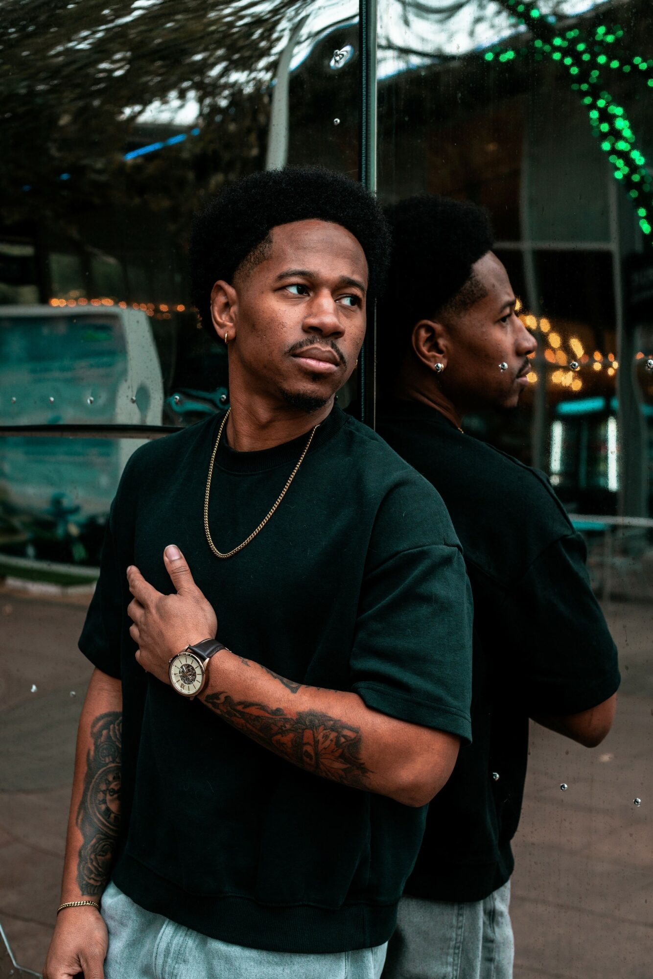 Two men standing outdoors, one facing forward and the other in profile, with trees and lights in the background.