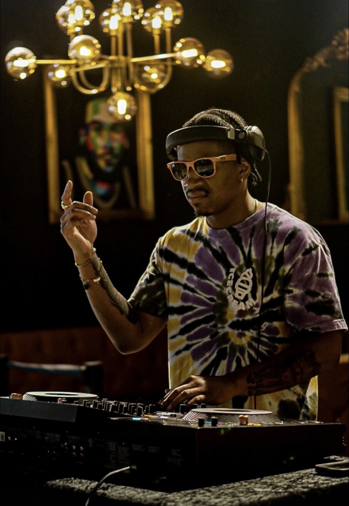 DJ wearing sunglasses and a tie-dye shirt, raising hand, in a dimly lit room with a portrait and chandelier in background.