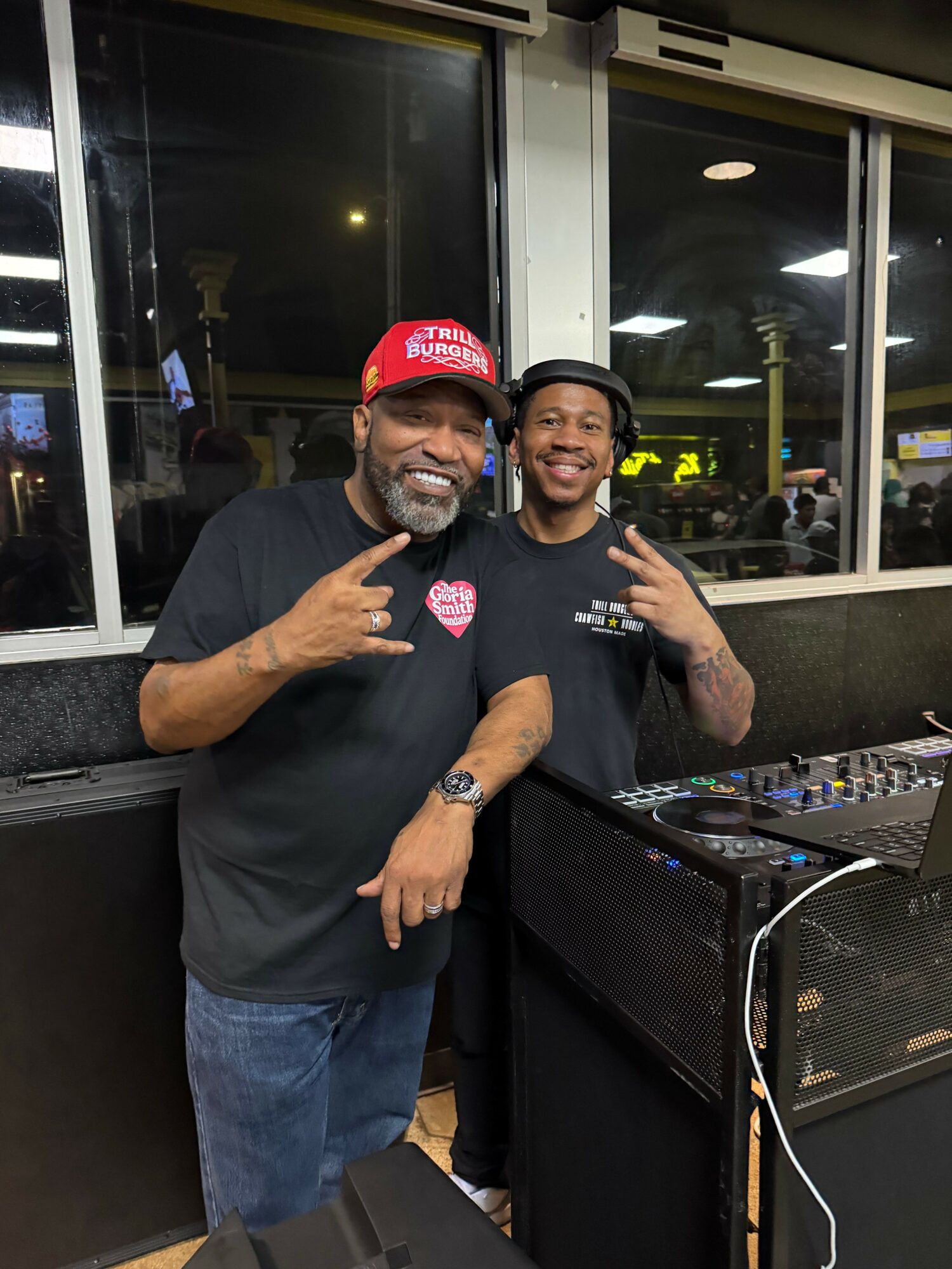 Two men in a room with DJ equipment, one wearing a red cap and glasses, the other making a hand gesture.