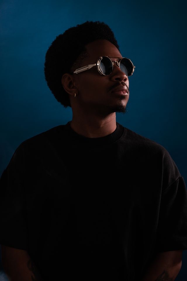 Person with short curly hair wearing sunglasses and a dark shirt, looking to the side against a dark background.