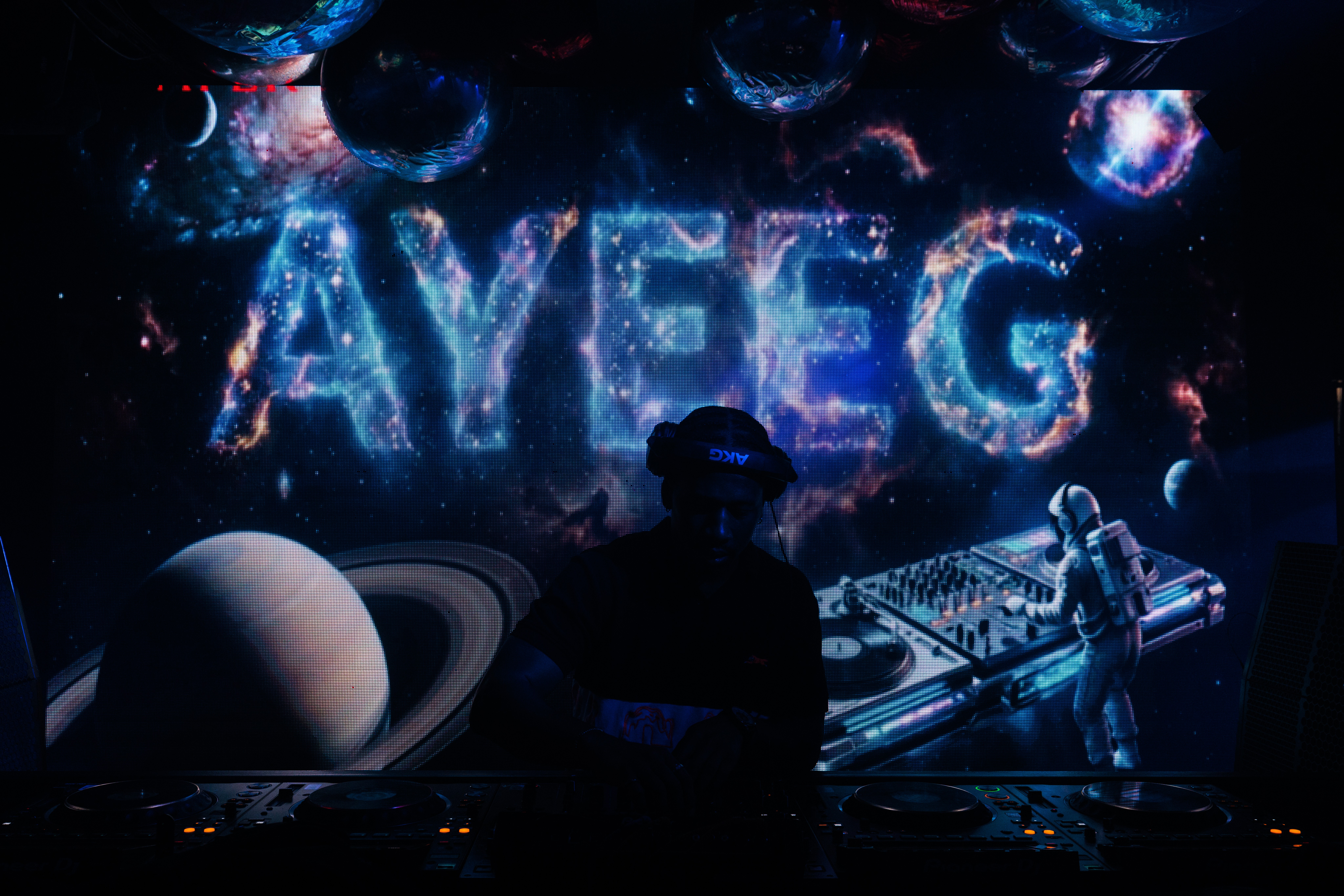 Dark scene with a DJ and a person in front of a large illuminated display reading 'AYE G', with planets and lights in the background.