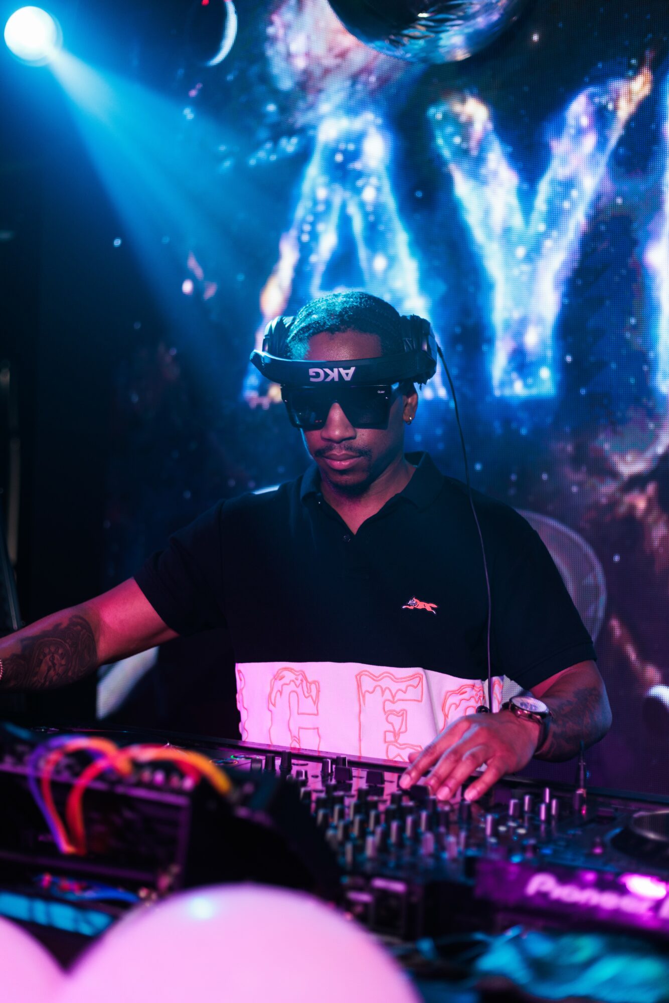 DJ wearing sunglasses and headphones, operating equipment in a dark club with colorful lights and a large illuminated 'AV' sign behind.