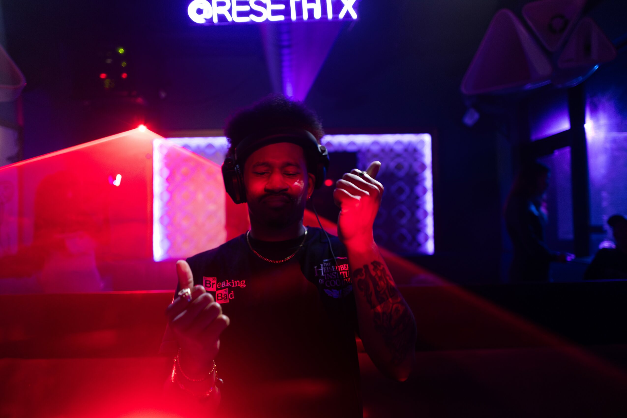 Person with headphones in a dimly lit club with colorful neon lights, smiling and holding a drink.