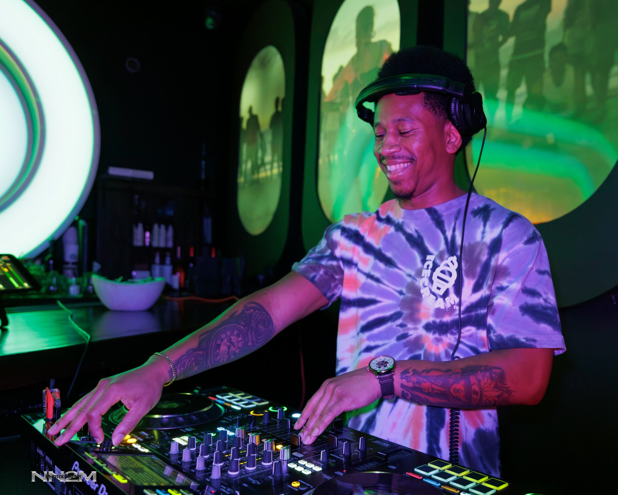 DJ smiling and mixing music at a club with colorful lights and screens in the background.