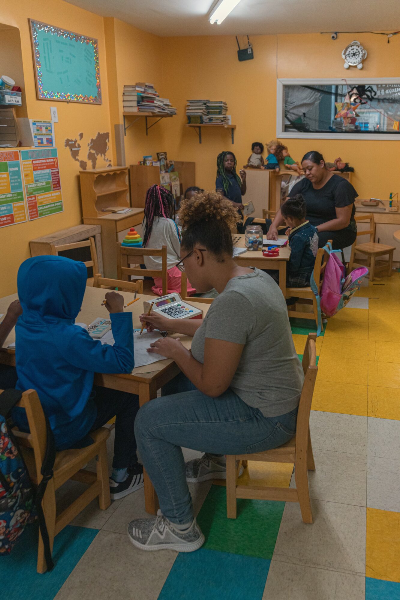 Classroom with children and teacher at desks, colorful walls, books, and educational materials, bright yellow floor, and a window.