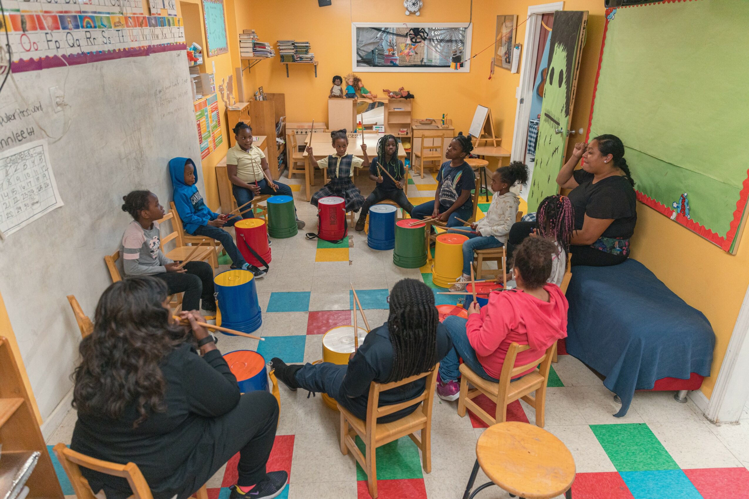 Children and teacher sitting in a circle in a colorful classroom with drums, chairs, and educational decorations.