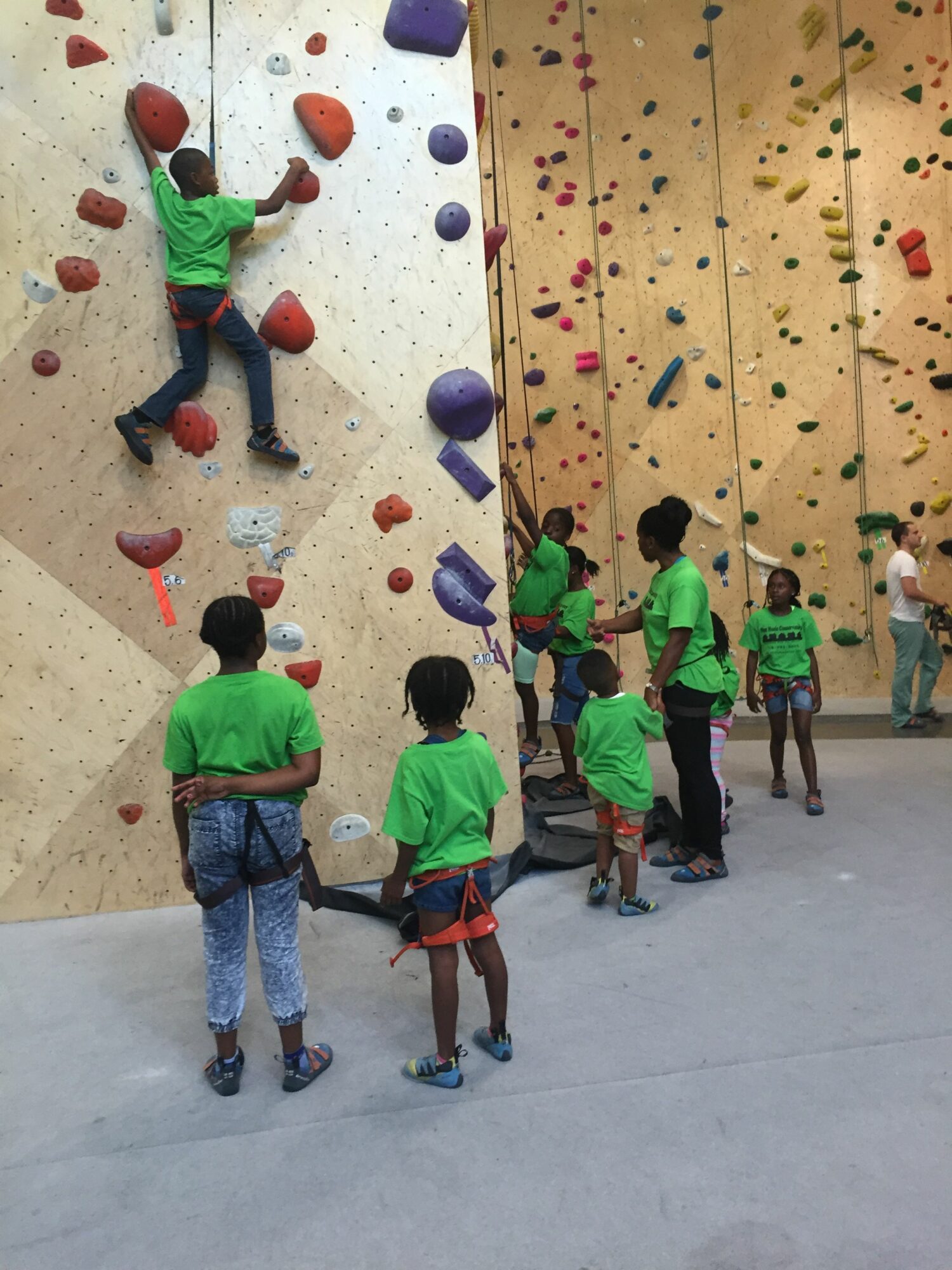 Children in green shirts are climbing and playing on an indoor rock climbing wall with colorful holds, in a gym.